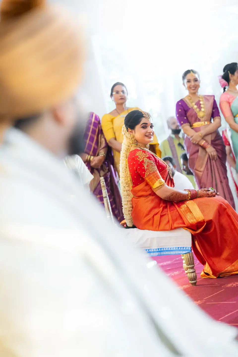 The bride seen past the groom's shoulder, seated among relatives in bright silks