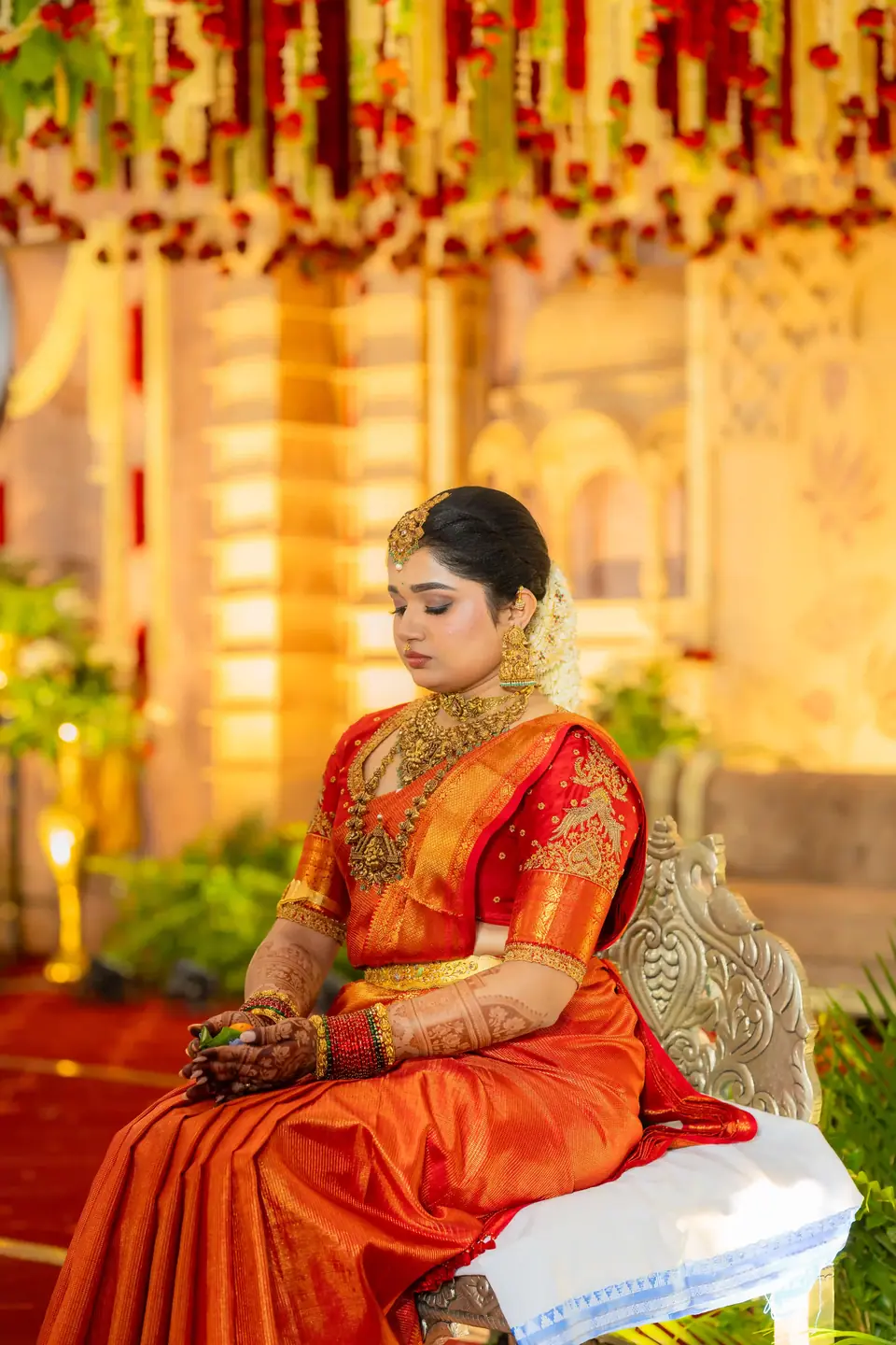 Bride in orange silk seated beneath hanging flowers, eyes lowered, hands resting in her lap