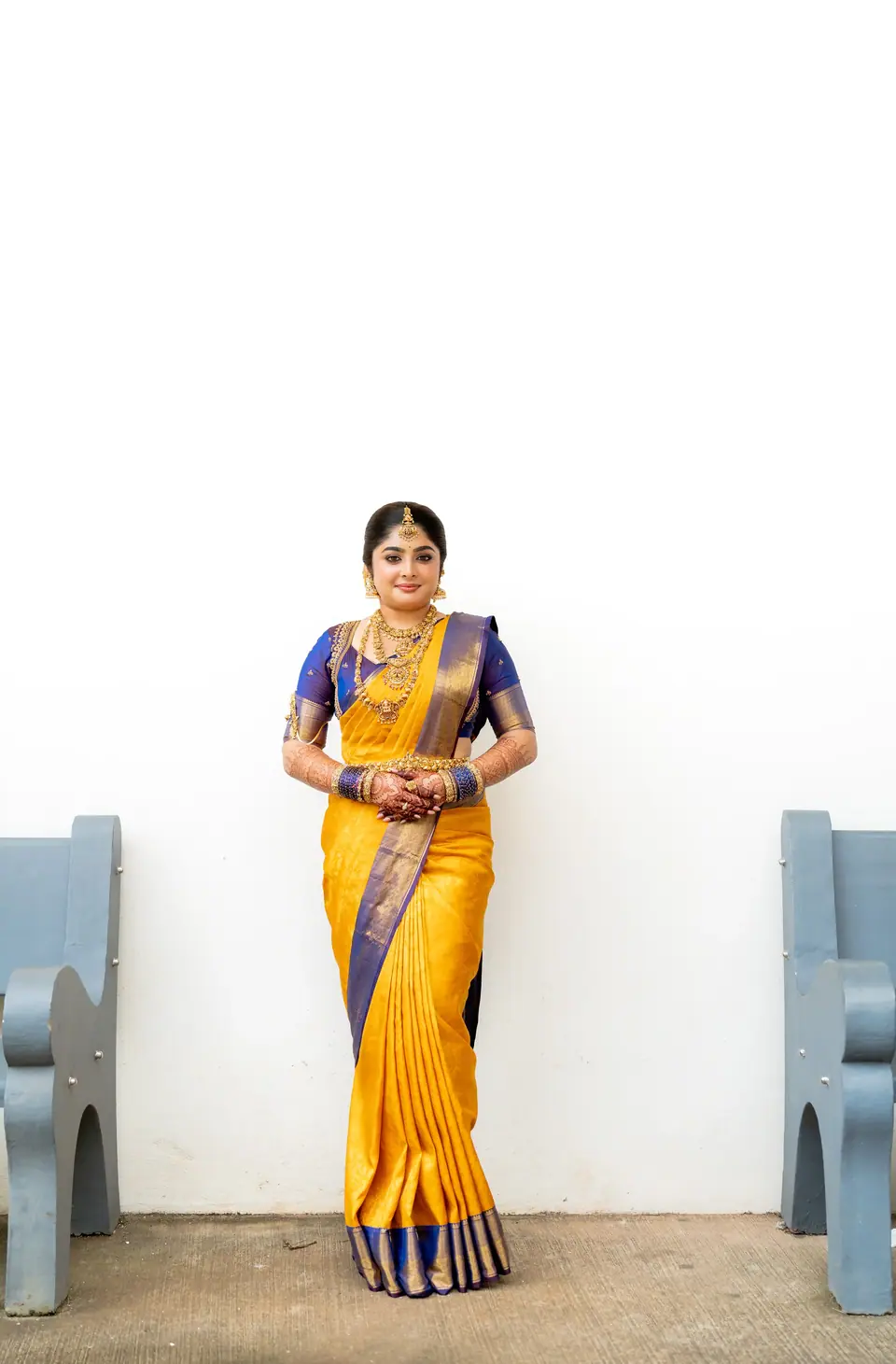 Bride in yellow and blue silk standing against a white wall between two grey benches