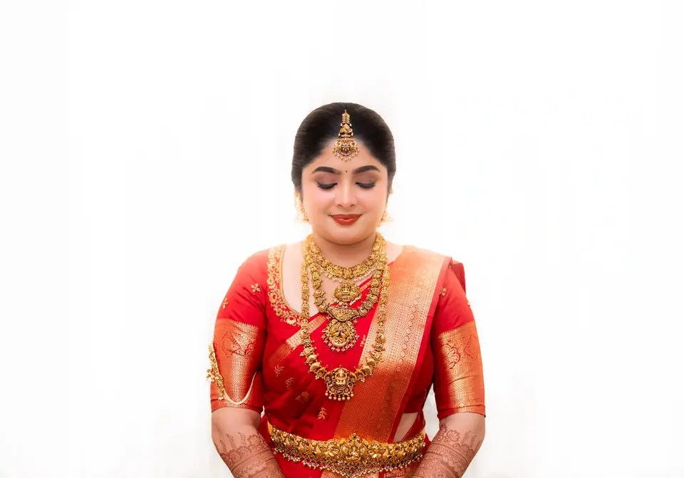 The bride in red silk seated against a bright white wall, eyes lowered