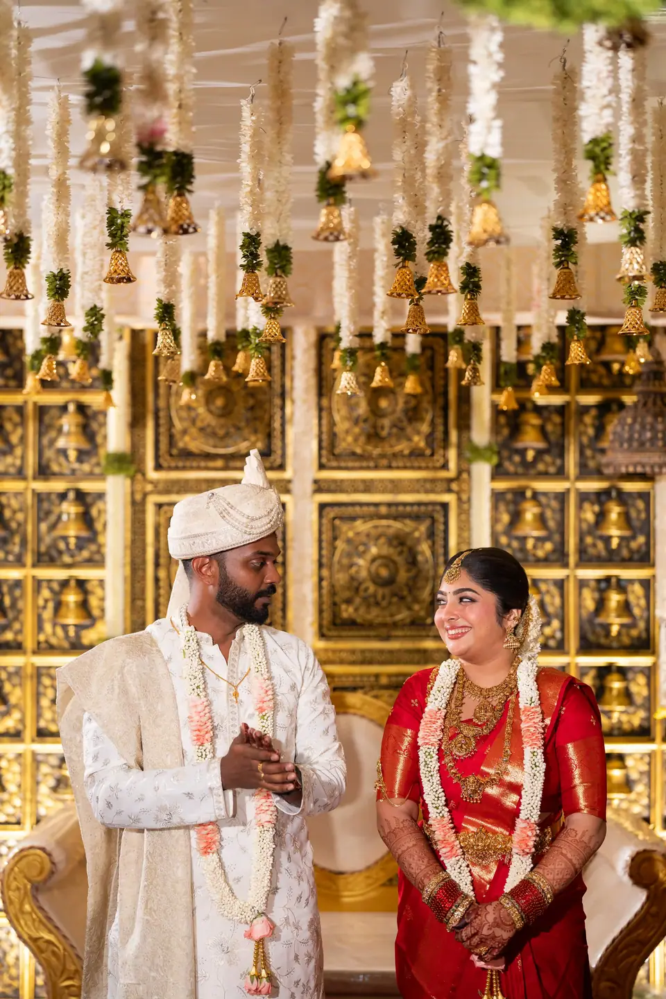 The couple standing before a carved gold backdrop, hands folded, smiling