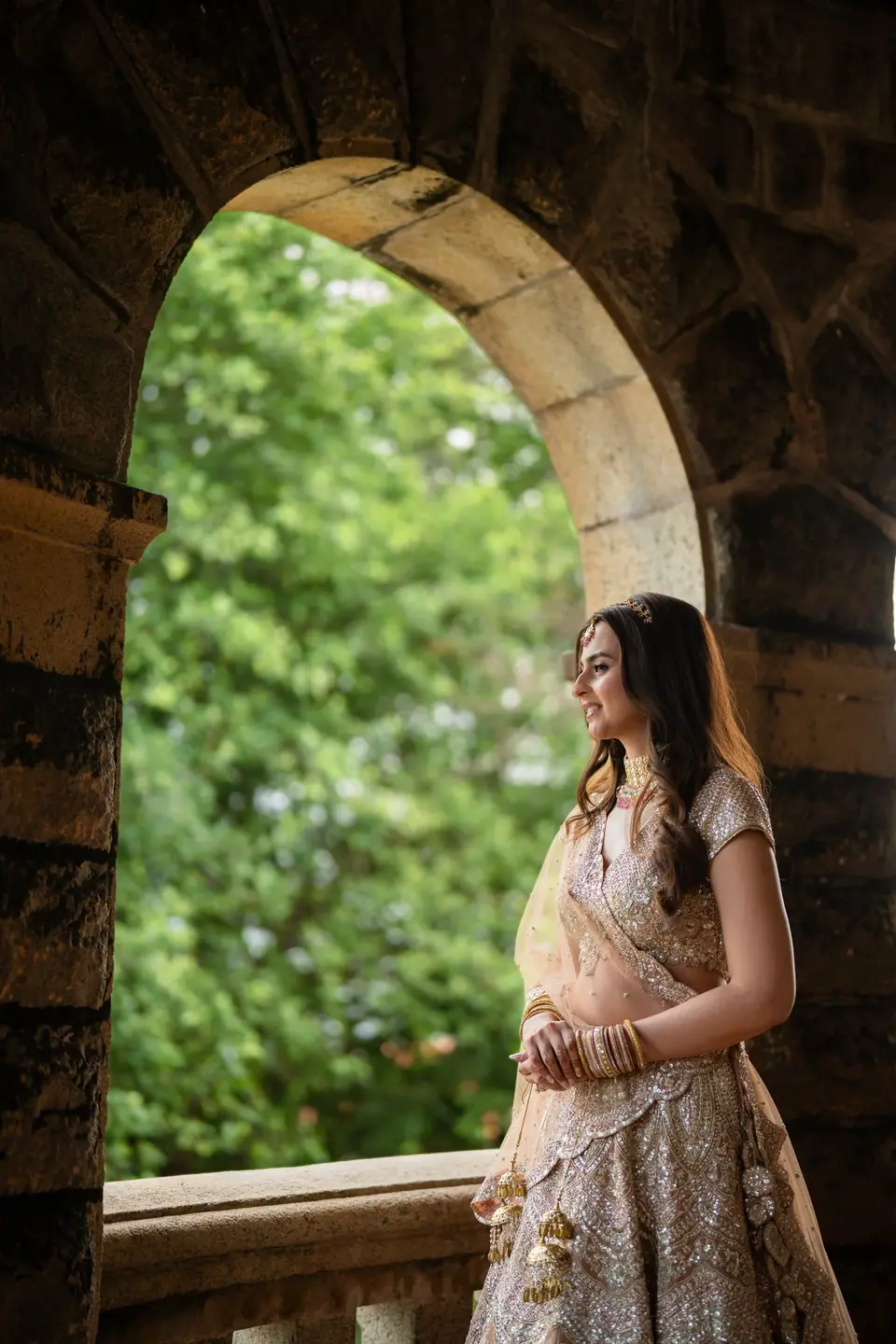The bride in a blush lehenga standing in a stone arch, green garden behind her