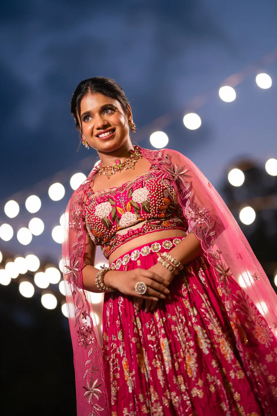 The bride in pink under the evening sky, a string of lights glowing behind