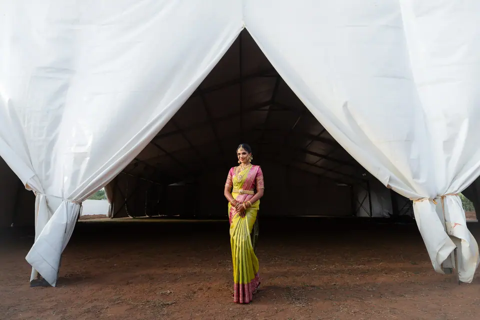 The bride standing alone at the opening of a vast white tent, curtains drawn back