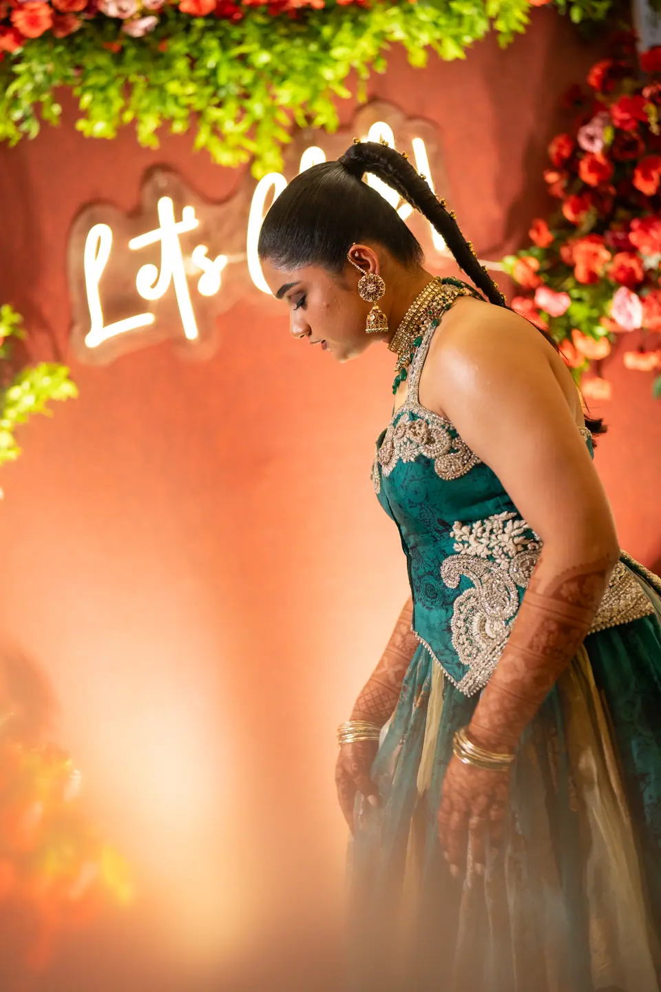 The bride looking down beside a neon sign and a wall of red roses